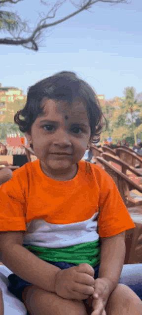 a little girl wearing an orange white and green shirt is sitting on someone 's lap