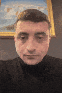 a man in a black turtleneck looks at the camera in front of a framed painting