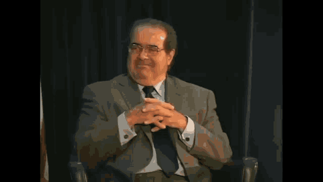 a man in a suit and tie is sitting in a chair with his hands folded