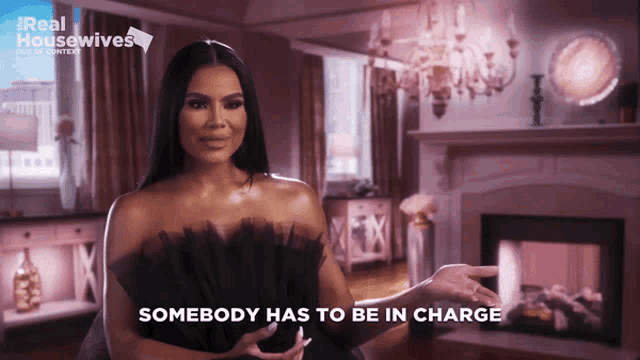a woman in a black dress says somebody has to be in charge in front of a fireplace