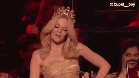 a woman in a gold dress and tiara is singing into a microphone on stage .