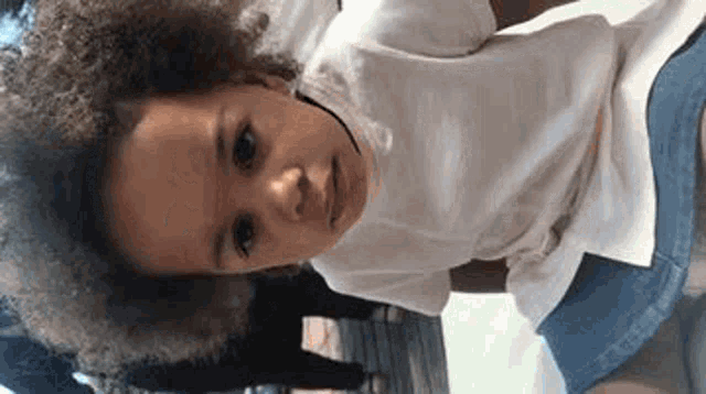 a little girl with curly hair and a white shirt is sitting on the ground .