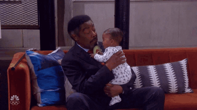 a man in a suit is holding a baby on a couch with the nbc logo