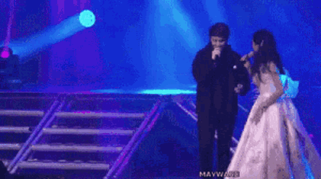 a woman in a white dress is singing into a microphone while standing next to a man in a black suit