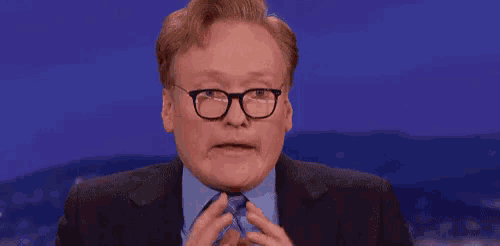 conan o'brien is wearing glasses and a suit and tie while making a funny face .