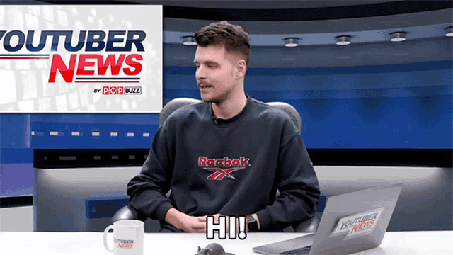 a man wearing a reebok sweatshirt sits in front of a youtuber news studio