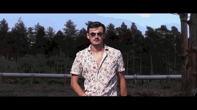 a man wearing sunglasses and a floral shirt stands in front of trees