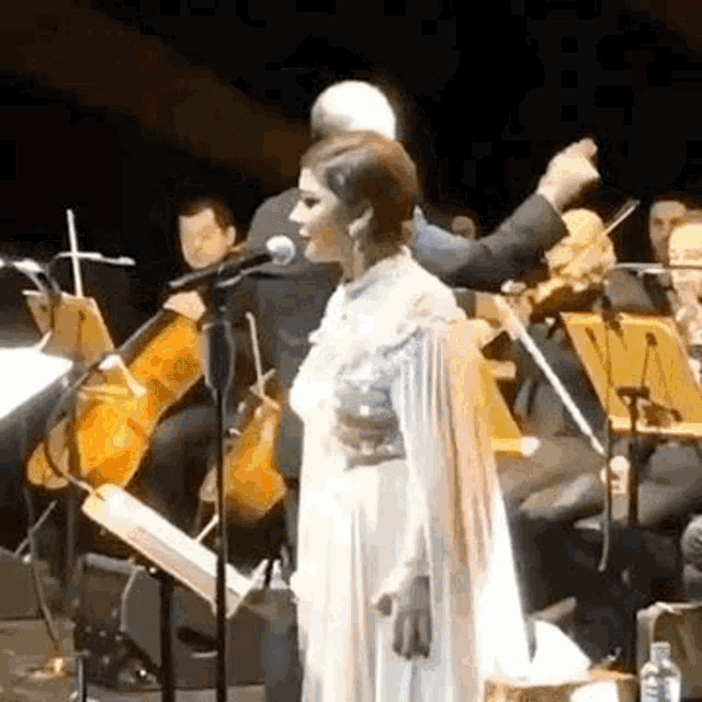 a woman is singing into a microphone on a stage with an orchestra in the background .