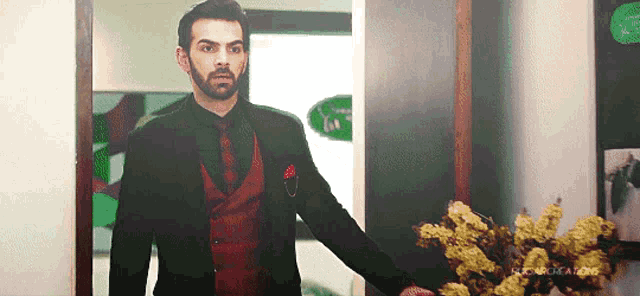 a man in a black suit and red vest is standing in front of a vase of flowers