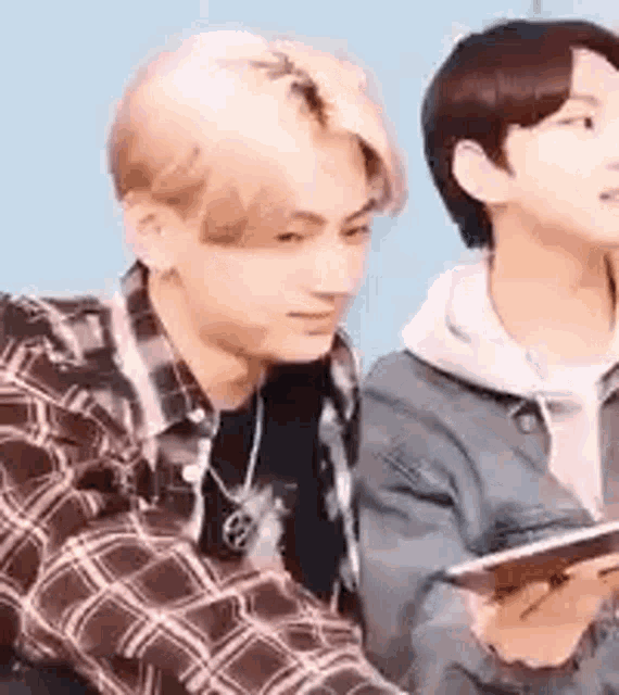 a man in a plaid shirt is sitting next to another man in a hoodie holding a cell phone .