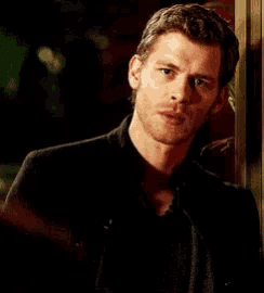 a man in a black shirt is standing in front of a door holding his hand to his chest .