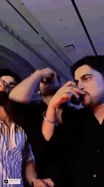 a man is drinking from a glass while standing next to another man in a dark room .
