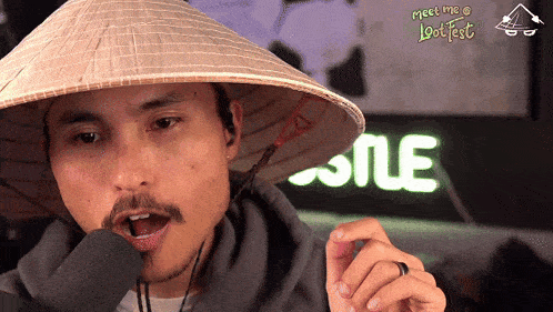 a man wearing a conical hat talks into a microphone with the words meet me @ lootfest in the background