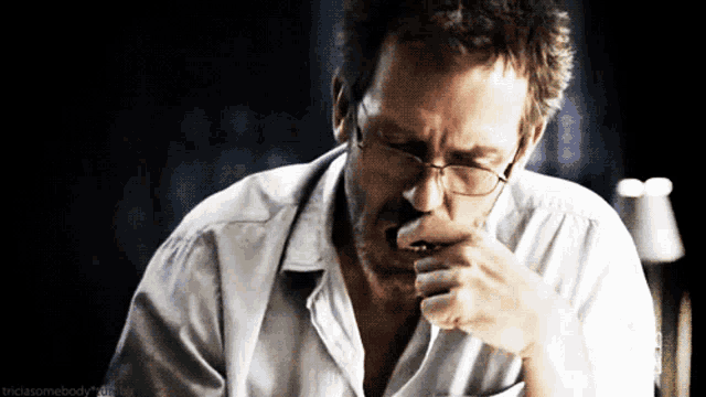 a man wearing glasses and a white shirt is coughing into his hand