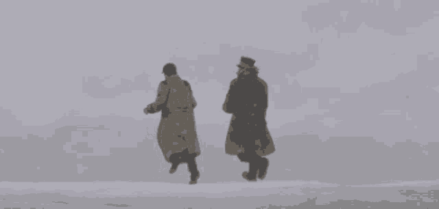 two men in trench coats and hats are walking through the snow .