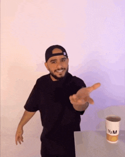 a man with a beard is standing in front of a cup of coffee and reaching out his hand .