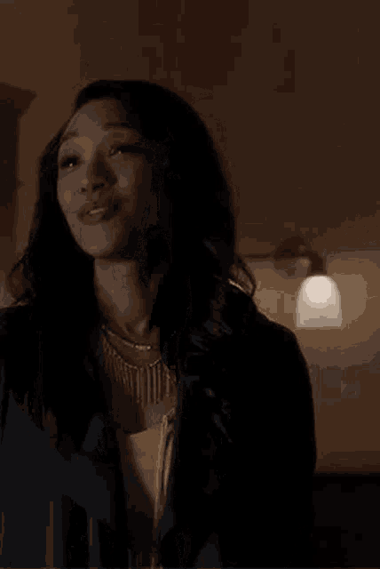 a woman is smiling in a dark room while wearing a black jacket and necklace .