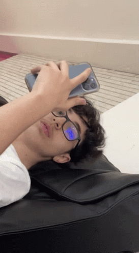 a young man wearing glasses is laying on a black pillow and looking at his phone