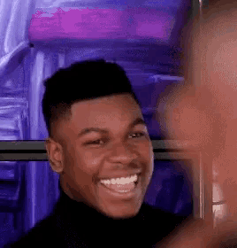 a young man in a black turtleneck is smiling while standing in front of a purple background .