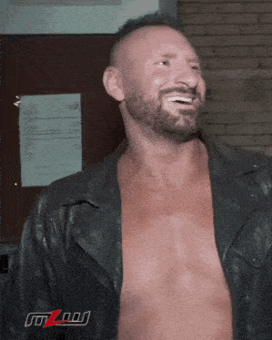 a shirtless man in a leather jacket is smiling in front of a wall with a sign that says mzn