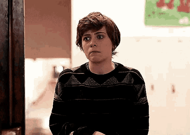 a young man in a sweater is standing in a hallway with his arms crossed and making a funny face .