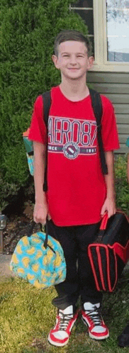 a young boy in a red shirt is standing in front of a house holding a backpack and lunch bag .