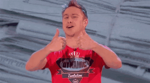 a man wearing a red shirt that says dynamite evolution gives two thumbs up