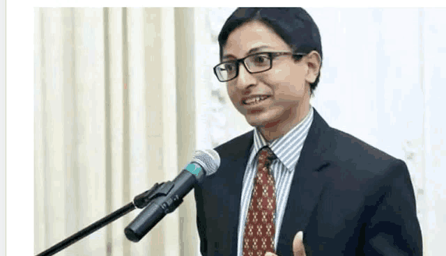the man is wearing glasses and a suit and tie and is speaking into a microphone .
