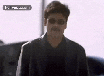 a man wearing sunglasses and a black jacket is walking towards a car .