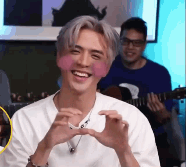 a man is smiling and making a heart with his hands while another man plays a guitar in the background .