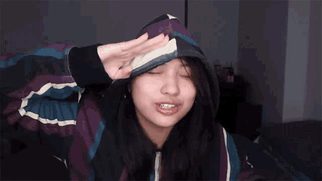 a woman wearing a purple and blue striped hoodie holds her hand to her forehead