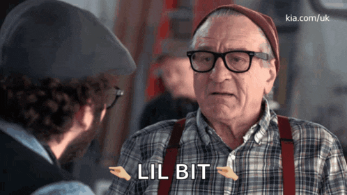 a man in a plaid shirt and suspenders is talking to another man and says lil bit