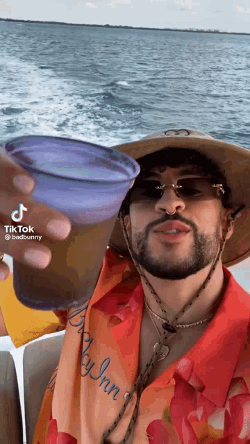 a man wearing a hat and sunglasses is holding a cup in front of a body of water .