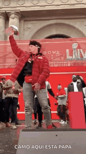 a man in a red jacket is standing on a stage in front of a crowd holding a football .