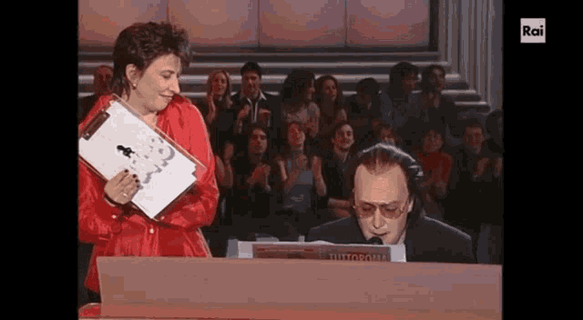 a woman in a red jacket is standing next to a man playing a piano with the word rai on the screen
