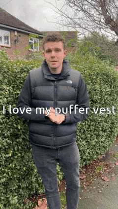 a man wearing a black puffer vest is standing in front of a hedge and says i love my puffer vest