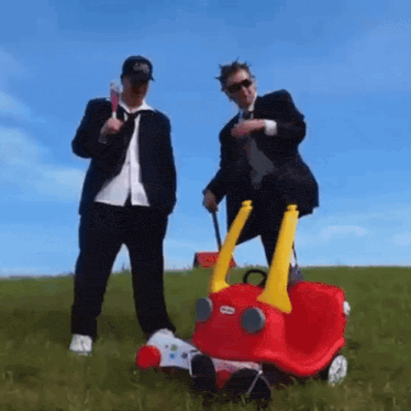 two men standing next to a red little tikes car