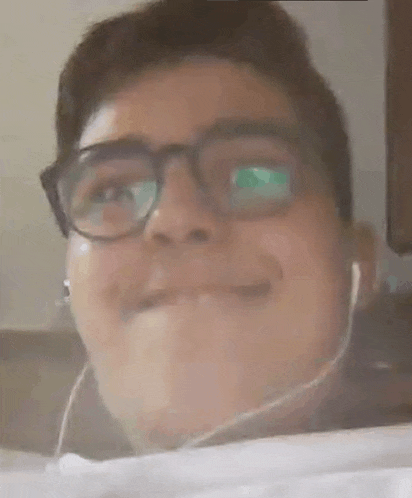 a young man wearing glasses and ear buds is smiling and making a funny face .