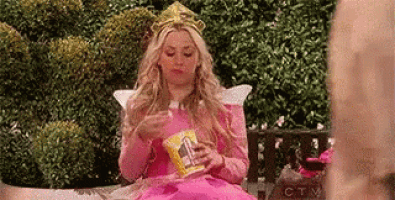 a woman in a pink dress and tiara is eating popcorn while sitting on a bench .