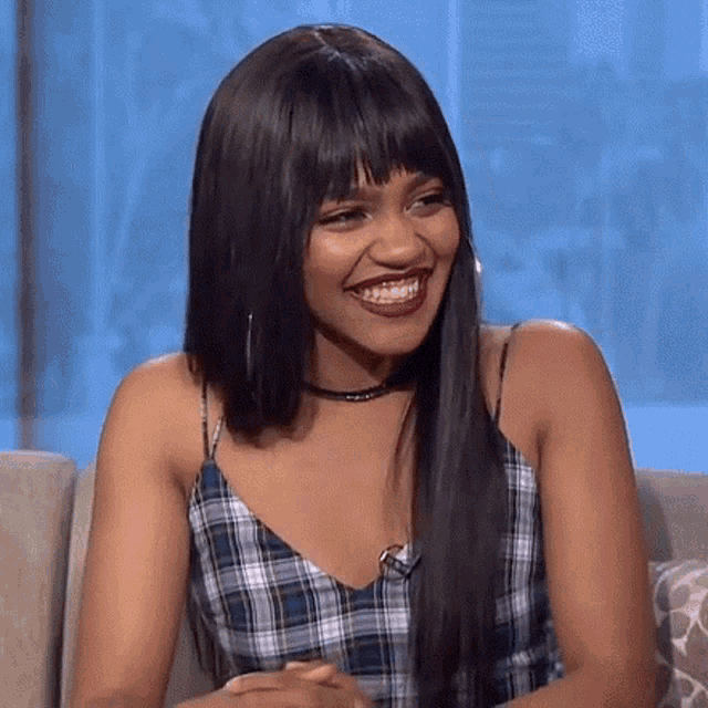 a woman is sitting on a couch smiling and wearing a plaid top .