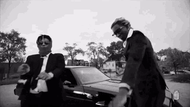 two men in suits and ties are standing next to each other in front of a car .