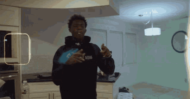 a man wearing a black hoodie with the number 6306 on it stands in a kitchen