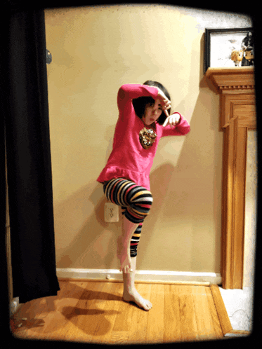 a young girl in a pink shirt and striped pants is standing on one leg