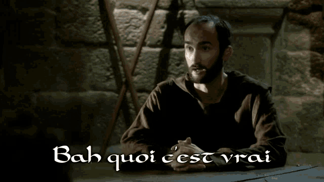 a man with a beard is sitting in front of a stone wall with the words bah quoi cest vrai written below him