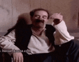 a man with a mustache is sitting in a chair smoking a cigarette and smiling .