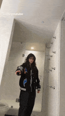 a girl in a black jacket is standing in a bathroom next to a toilet and a shower .