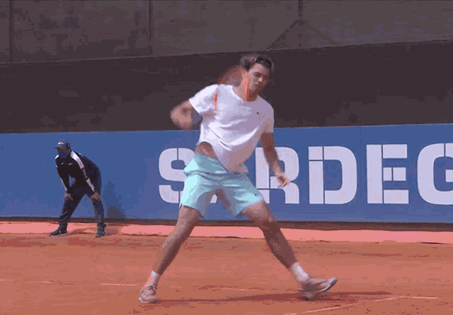 a man in a white shirt and blue shorts is playing tennis in front of a sign that says sardeg