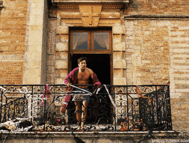 a shirtless man is standing on a balcony with a wrought iron railing and a brick building in the background