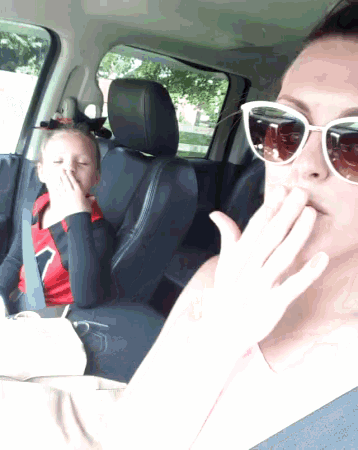a woman wearing sunglasses covers her mouth while a little girl sits in the back seat