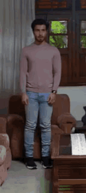 a man in a pink sweater and jeans is standing in a living room next to a couch .